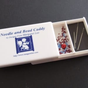 May include: White plastic needle and bead caddy with a blue and white logo that says "Needle and Bead Caddy by Designs from Margaret Lee http://www.margaretien.com". The caddy has two compartments, one with a red and blue bead assortment and the other with silver needles.