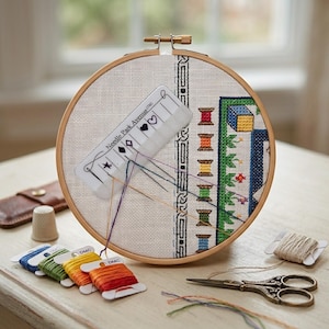 May include: A round wooden embroidery hoop holds white Aida fabric with a partially completed cross-stitch design. A Needle Park Avenue thread organizer is attached, featuring colorful embroidery floss, a metal thimble, and scissors.