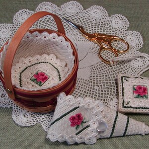 Puede incluir: Una cesta de crochet blanca con un asa de madera, llena de manteles individuales de crochet blanco y un pequeño bordado de rosa roja. Un cojín de alfileres de crochet blanco con un borde verde y un bordado de rosa roja. Un estuche de agujas de crochet blanco con un borde verde y un bordado de rosa roja. Un par de tijeras doradas con un mantel individual de crochet blanco.