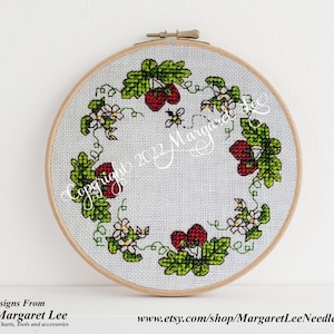 Puede incluir: Un aro de bordado de punto de cruz con fondo blanco y una corona de hojas verdes, fresas rojas y flores blancas. Una abeja vuela cerca de las fresas. El texto "Copyright © Margaret Lee" es visible en la esquina inferior izquierda del aro.