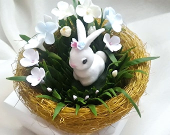May include: A white bunny figurine sits in a gold nest with white and blue flowers and green grass.