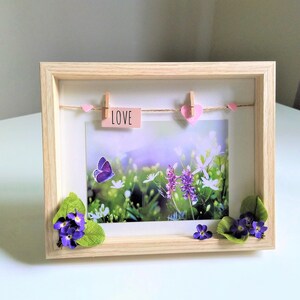 May include: A wooden picture frame with a nature scene print. The frame features a string with pink hearts and a small sign that says "LOVE". A purple butterfly and flowers are on the print, with additional flowers and leaves attached to the frame.