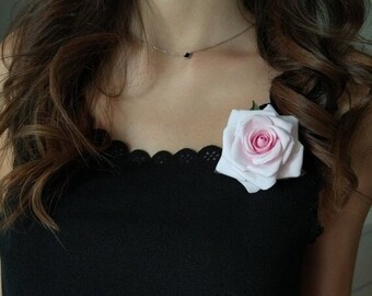 May include: A black top with a scalloped neckline, adorned with a light pink rose with a darker pink center. A delicate silver necklace with a small black stone is also visible. The word "Nelyn" is written in cursive.