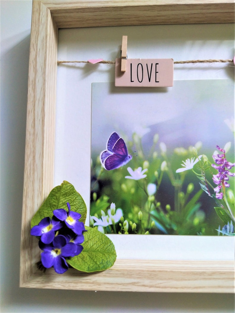 May include: A light wood shadow box frame containing a photograph of a purple butterfly and white flowers. A small wooden clothespin holds a pink tag with the word LOVE printed in black. Artificial purple flowers and green leaves are attached.