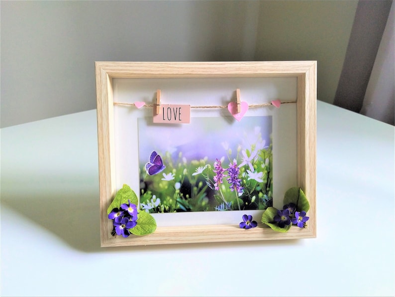 May include: A wooden picture frame with a nature scene print. The frame features a string with pink hearts and a small sign that says "LOVE". A purple butterfly and flowers are on the print, with additional flowers and leaves attached to the frame.