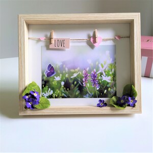 May include: A framed picture with a floral scene, a butterfly, and the word "LOVE". The frame is light wood, with decorative flowers and leaves. Pink hearts and clothespins adorn a string above the picture.