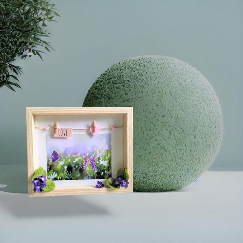 May include: A light wood shadow box frame containing a photograph of a field with a purple butterfly and flowers. A string with a pink tag reading LOVE and two small pink heart clips hangs inside. Purple silk flowers decorate the bottom corners.