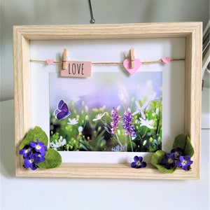 May include: A wooden picture frame with a nature scene inside. The frame has a string with clothespins holding pink hearts and a card that says "LOVE". The picture shows a butterfly and purple and white flowers.