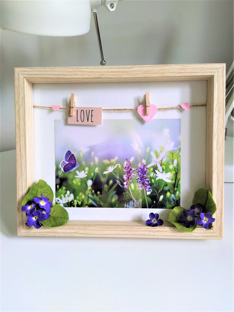 May include: A wooden picture frame with a nature scene inside. The frame has a string with clothespins holding pink hearts and a card that says "LOVE". The picture shows a butterfly and purple and white flowers.