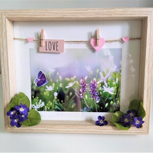 May include: A wooden picture frame with a white mat and a photo of a field of flowers and a butterfly. The photo is decorated with pink hearts and a pink tag that says "LOVE".