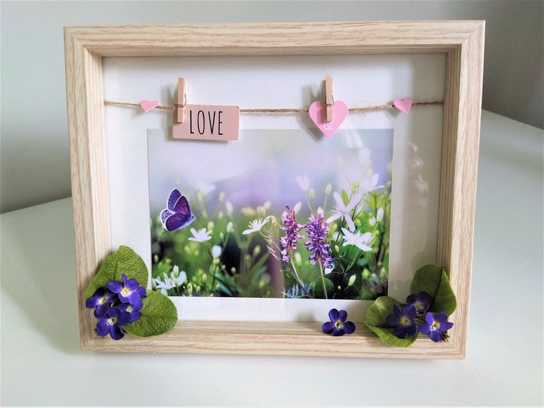 May include: A wooden picture frame with a white mat and a photo of a field of flowers and a butterfly. The photo is decorated with pink hearts and a pink tag that says "LOVE".