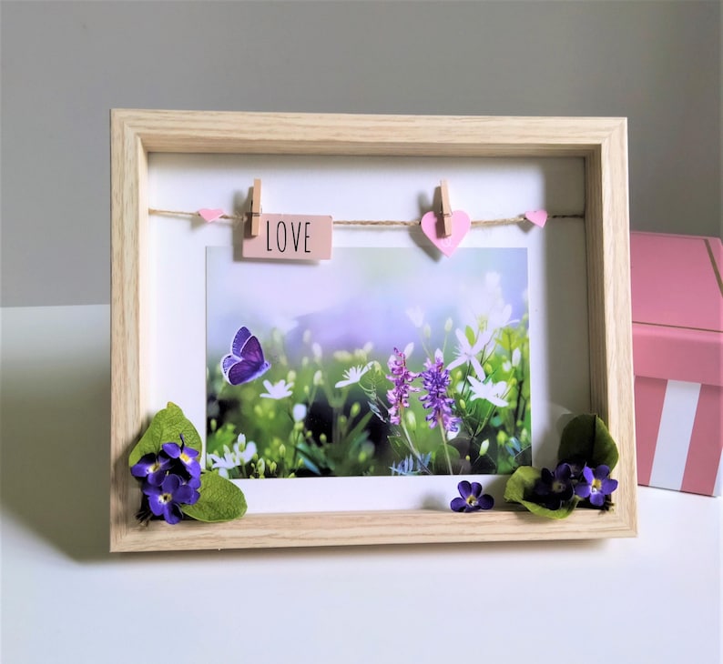 May include: A framed picture with a nature scene, featuring a purple butterfly and flowers. The wooden frame holds a photo with a string and small pink hearts. A wooden sign with the word "LOVE" is attached. Artificial flowers are placed at the bottom.