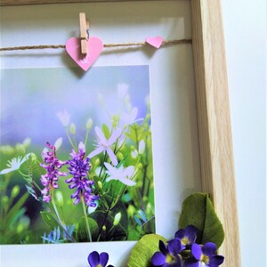 May include: A light wood shadow box frame containing a photograph of purple and white wildflowers. A jute twine string is stretched across the top with a pink heart-shaped clip and a small pink heart. Artificial purple flowers are at the bottom.