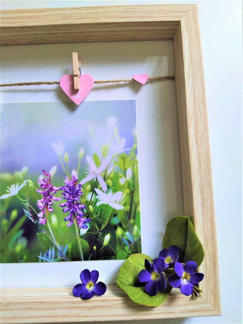 May include: A light wood shadow box frame containing a photograph of purple and white wildflowers. A jute twine string is stretched across the top with a pink heart-shaped clip and a small pink heart. Artificial purple flowers are at the bottom.