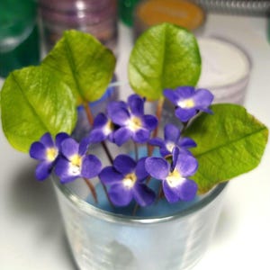 May include: A small glass filled with artificial violets. The flowers are a deep purple with yellow centers, and the leaves are a vibrant green. The arrangement is set against a blurred background of colorful containers.
