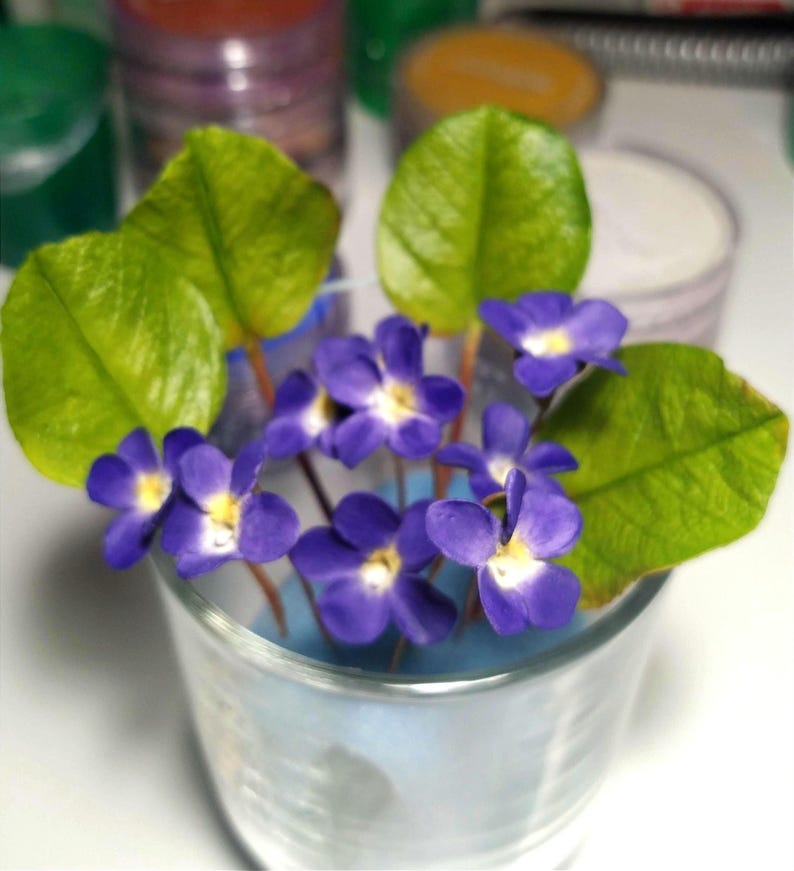 May include: A small glass filled with artificial violets. The flowers are a deep purple with yellow centers, and the leaves are a vibrant green. The arrangement is set against a blurred background of colorful containers.