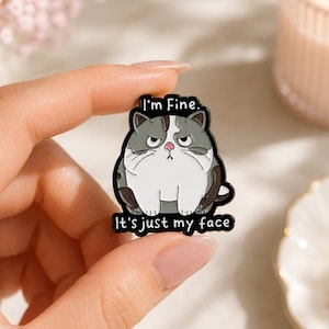 May include: A black and white enamel pin featuring a grumpy grey and white cat with a pink nose. The text "I'm Fine." is printed in white above the cat, and "It's just my face" is printed in white below the cat on a black background.