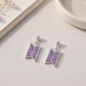 May include: A pair of silver-toned stud earrings featuring a trapezoidal pendant design. Each pendant is inlaid with numerous small, round, purple-colored stones arranged in a grid pattern, suspended from a simple silver ball stud post.