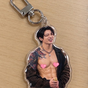 May include: A clear acrylic keychain featuring a printed image of a person with dark hair, a silver chain necklace, and a dark jacket worn off the shoulders. The person has visible arm tattoos and two pink heart-shaped stickers covering the chest area.