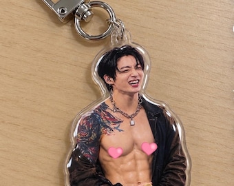 May include: A clear acrylic keychain featuring a printed image of a person with dark hair, a silver chain necklace, and a dark jacket worn off the shoulders. The person has visible arm tattoos and two pink heart-shaped stickers covering the chest area.