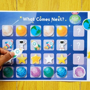 Puede incluir: Un juego educativo con temática espacial con el texto "What Comes Next?" presenta planetas, cohetes, astronautas y estrellas. Una mano coloca una pegatina de cohete en el tablero de juego. El juego está sobre un fondo azul.