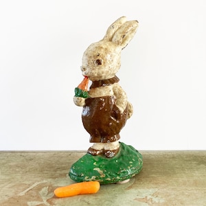 May include: A vintage cast metal rabbit figurine with a distressed off-white finish, wearing brown trousers and a dark collar. The rabbit holds a small orange carrot with green leaves in its paw, standing on a textured green base with a loose carrot.