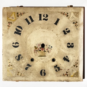 May include: A white wooden clock face with black Roman numeral hour markers and a floral design in the corners. The clock face has a small hole in the center and a painted floral design below it.