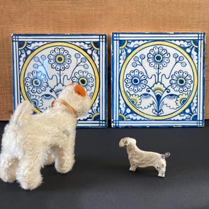 May include: Two square ceramic tiles with blue and yellow floral designs. A white stuffed dog with an orange ear and a small white dog figurine are in front of the tiles. The tiles have a blue border.