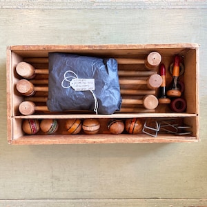May include: A wooden box filled with a vintage croquet set. The set includes wooden mallets, balls, and wickets. The box is labeled "Croquet Set 1912, Made in England,  Woodbridge, 1912."