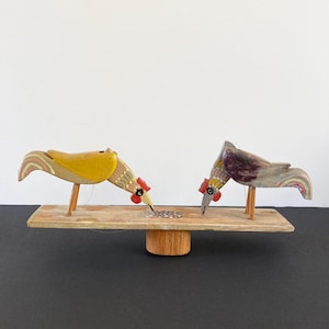 May include: Two wooden toy birds painted in yellow, gray, and red are perched on a wooden platform. The birds are facing each other and appear to be pecking at a pile of seeds in the center of the platform.