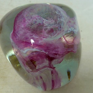 May include: A clear glass paperweight with a swirling pink and white design inside. The paperweight is round and has a smooth, polished surface.