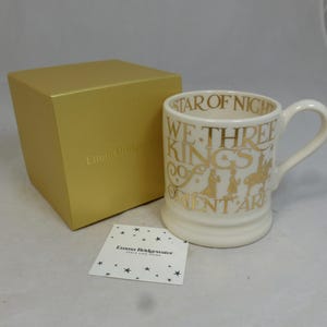 Può includere: Una tazza in ceramica color crema con scritte e illustrazioni dorate, con il testo "STAR OF NIGHT WE THREE KINGS OF ORIENT ARE". Sono presenti anche una scatola dorata e una piccola carta con grafiche a stella.