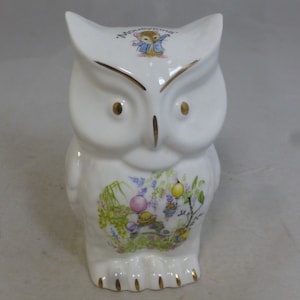 May include: White ceramic owl money bank with a gold trim. The owl has a floral design on its body and a small image of Peter Rabbit on its head. The text "Peter Rabbit" is written in gold on the owl's head.