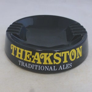 Puede incluir: Cenicero de cerámica negro con un logotipo amarillo "Theakston Traditional Ales".