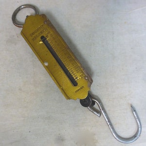 Peut inclure: Une balance à suspendre vintage de couleur dorée avec une aiguille noire et un crochet en bas. L'échelle est marquée de nombres de 0 à 25. La balance est étiquetée "Pocket Balance" et "Foreign".