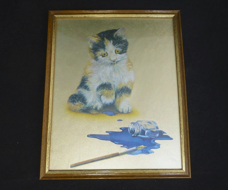Puede incluir: Una impresi&oacute;n enmarcada de un gatito calico sentado junto a un frasco de pintura azul derramado y un pincel. El fondo es dorado.