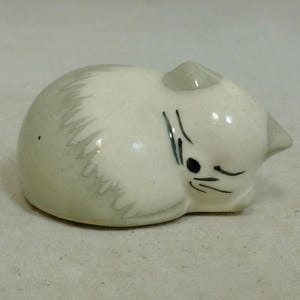 May include: A small, ceramic figurine of a curled-up cat. The cat is primarily white with gray accents on its back and ears. Black lines depict the eyes and whiskers. The figurine has a glossy finish.
