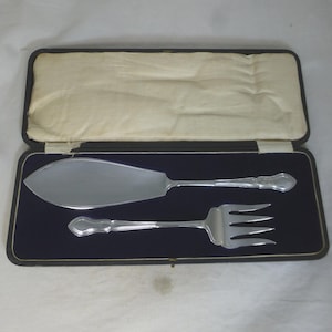 May include: A silver cake server and fork set in a black velvet lined box. The set is made of polished metal and has a traditional design.