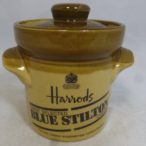 May include: A brown ceramic jar with a lid, featuring the Harrods logo and the text "Finest Selected Blue Stilton Cheese". The jar has two handles on the sides.