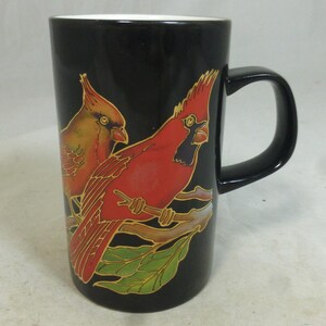 May include: Black ceramic mug featuring a vibrant illustration of two cardinals perched on a branch with green leaves. The birds are depicted in shades of red, orange, and yellow, with intricate details.