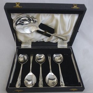 Puede incluir: Un juego de seis cucharas de plata y una cuchara de servir de plata en una caja forrada de terciopelo negro. Las cucharas están todas pulidas y brillantes.