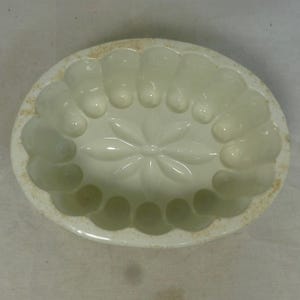 May include: An off-white, oval-shaped ceramic mold with a floral design in the center. The mold has a scalloped edge and is likely used for baking or creating decorative desserts. The interior features a detailed flower pattern.
