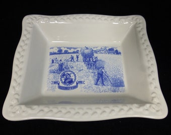 May include: A white ceramic serving dish with a blue and white design featuring a scene of farmers harvesting wheat. The dish has a raised border with a braided pattern. The text "1897 Shredded Wheat 1992" is printed on the dish.