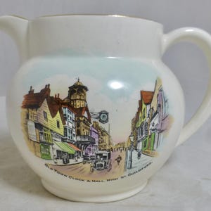 Può includere: Brocca in ceramica color crema con una dettagliata illustrazione di una scena di strada. L'immagine presenta edifici, una torre dell'orologio e il testo "Old Town Clock & Hall High St. Guildford". La brocca ha un beccuccio e un manico curvo.