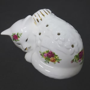 Puede incluir: Figura de gato de porcelana blanca en posición de dormir, adornada con calcomanías florales de rosas en tonos rosa, amarillo y rojo. La espalda del gato presenta detalles dorados y varios agujeros pequeños. Un artículo decorativo.