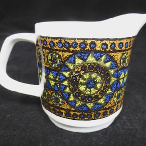 May include: A white ceramic creamer with a geometric pattern in shades of blue, yellow, and brown. The pattern features circles, triangles, and stars.