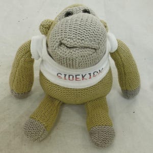 May include: A knitted monkey toy with a grey head and tan body. The monkey is wearing a white shirt with the word "SIDEKICK" in red and black letters. The toy has grey feet and hands.