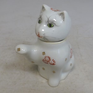 Peut inclure: Figurines de chat en porcelaine blanche avec des accents floraux rouges et dorés. Le chat est assis, la patte levée, et a un couvercle amovible en forme de tête de chat.