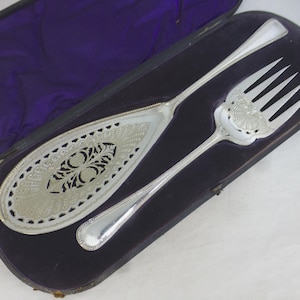 Puede incluir: Un juego de servicio de plata con un diseño ornamentado, que incluye una cuchara ranurada y un tenedor, en un estuche forrado de terciopelo morado.