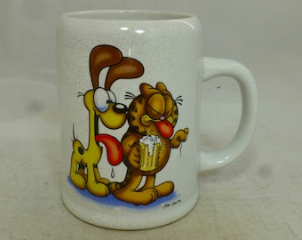 May include: White ceramic mug with a cracked glaze finish. The mug features a cartoon image of Garfield the cat and Odie the dog. Garfield is holding a beer mug and Odie is sticking out his tongue. The mug has the text "JIM DAVIS" at the bottom.